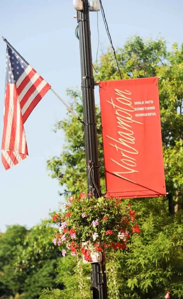 Closeup of a light pole in Northampton, MA with a flower basket, American flag, and a red flag with "Northampton: Walk into something wonderful" written on it.
