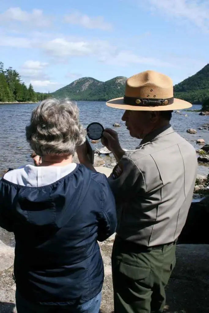 A man in a uniform shows a camera filter to a woman while overlooking the Bubble Mountains in Bar Harbor, Maine.