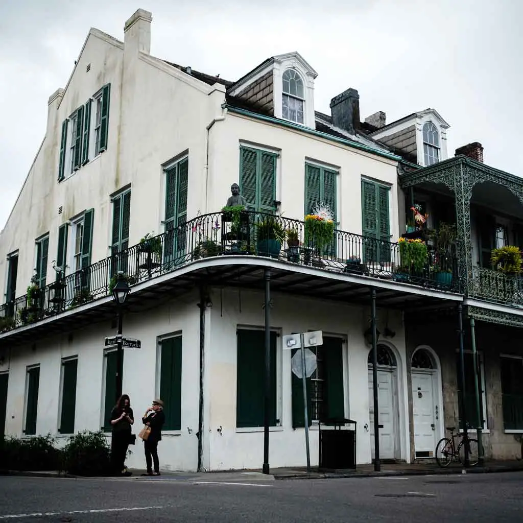 Close up of an old white house with green shutters and a decorative iron balcony; 2 women stand below on the sidewalk.