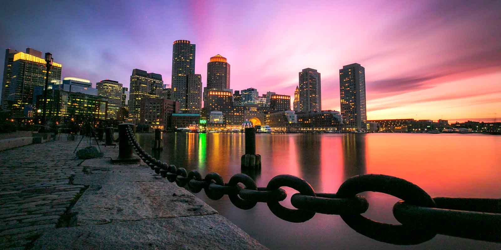 Landscape view of the Boston waterfront during sunrise with blue, purple, pink, yellow and orange skies with the cityscape in the background.