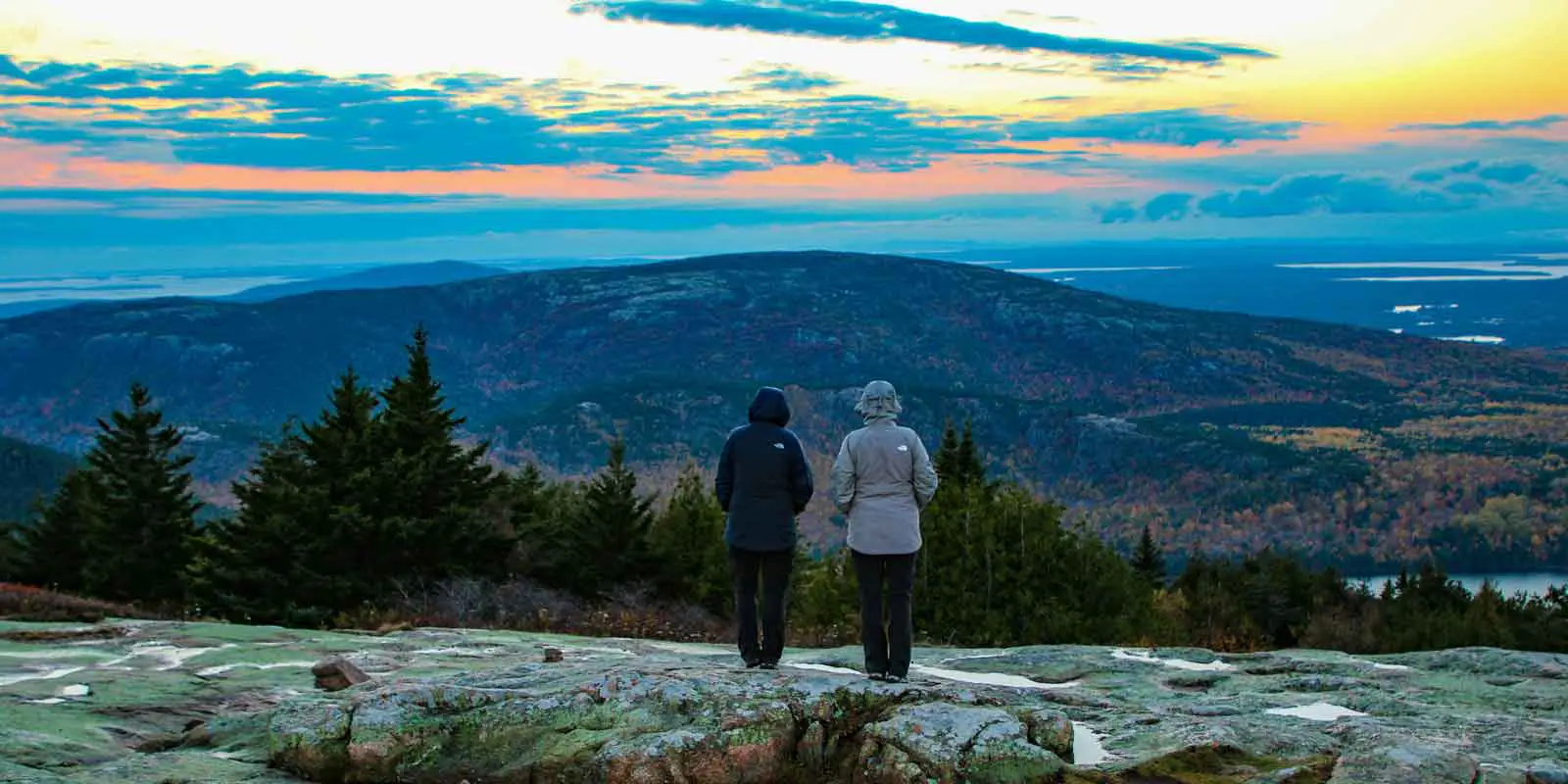 Two people in hooded jackets watching the sunset from Cadillac Mountain.