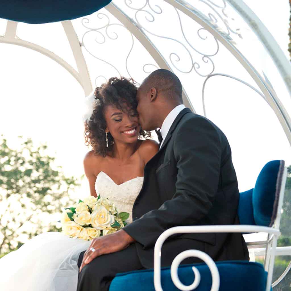 A groom kisses his smiling bride on the cheek while sitting in a pumpking-shaped carriage coach.