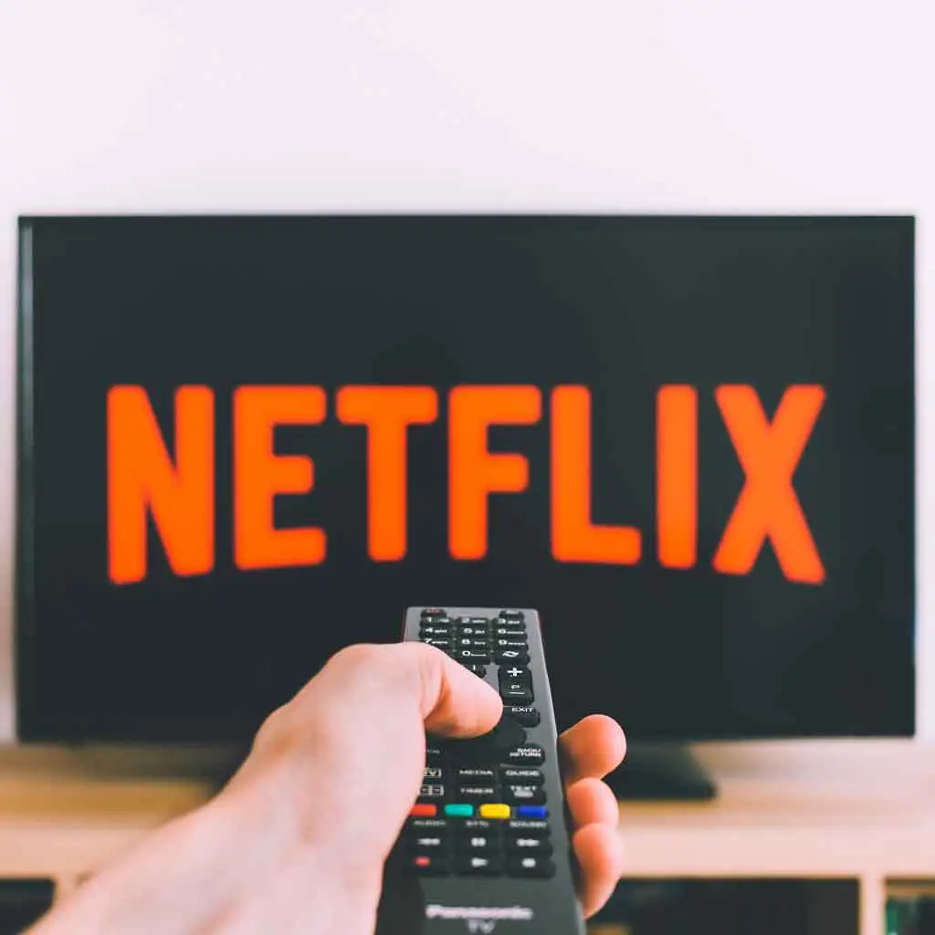 Closeup of a hand holding a remote pointing at a TV with the Netflix logo.
