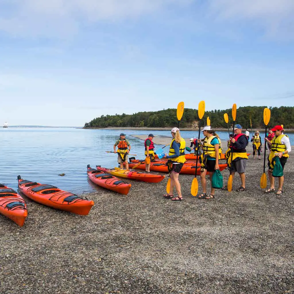 Photo of a group of people on a shoreline wearing life jackets and carrying oars with tandem kayaks nearby.