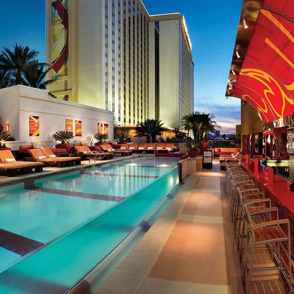 Landscape view of a pool deck with lounge chairs lined up on one side and a bar on the other side.