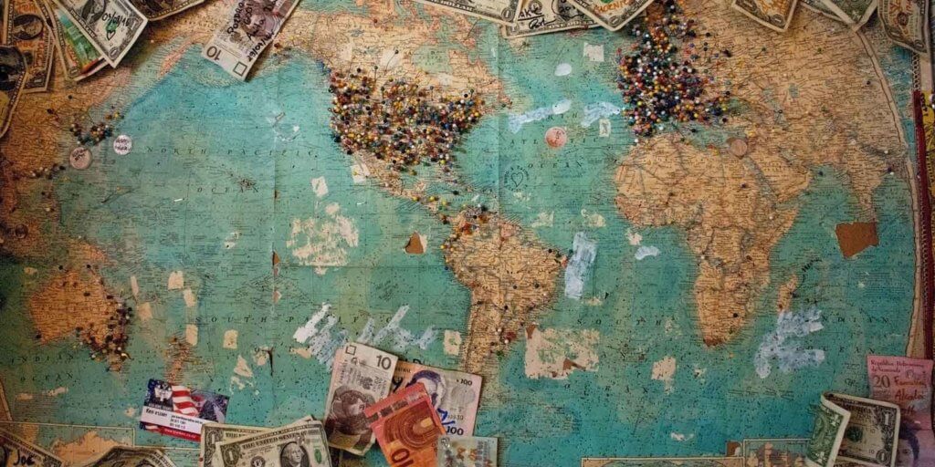 A map hung up on corkboard with lots of pins in different locations and currency from around the world attached.