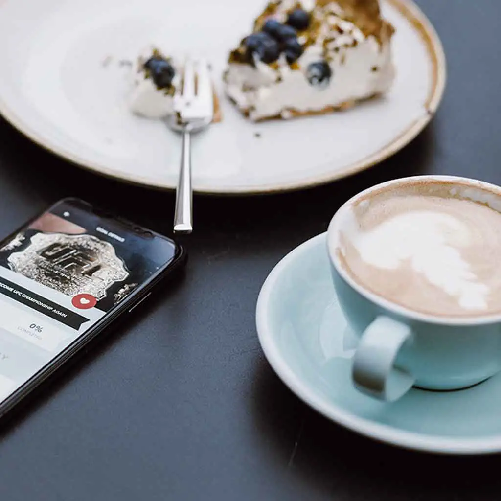 Closeup of a smartphone, plate with a half eaten piece of pie, and a cup filled with a latte.