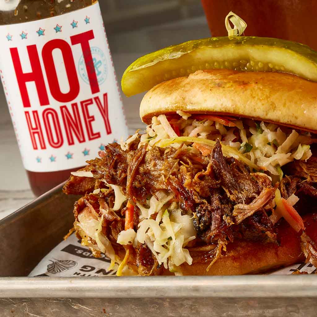 Closeup photo of a pulled pork sandwich with a bottle of hot honey in the background.