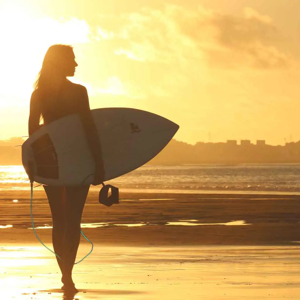 Silhouette of a woman carrying a surfboard with the sun shining on her.