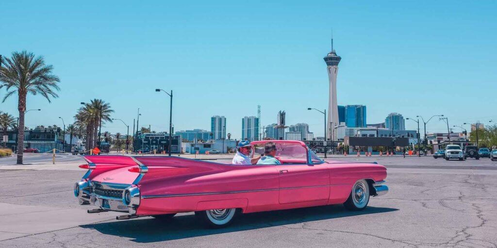 Two men riding in a pink Cadillac convertible in a city.