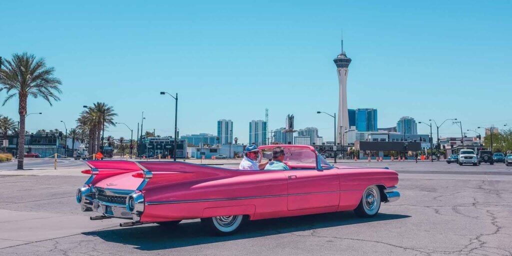 Two men riding in a pink Cadillac convertible in a city.