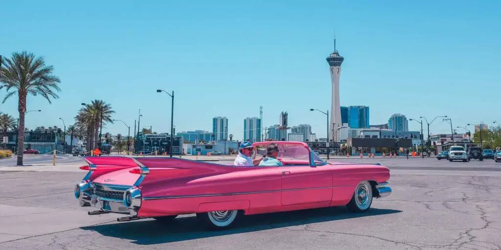Two men riding in a pink Cadillac convertible in a city.