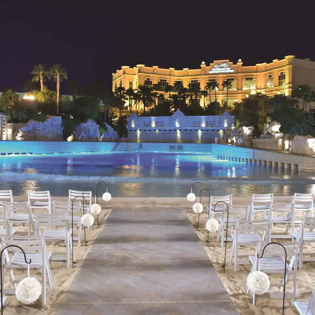 Landscape view of a wedding ceremony set up on a faux sand beach in front of a pool with a hotel in the background.