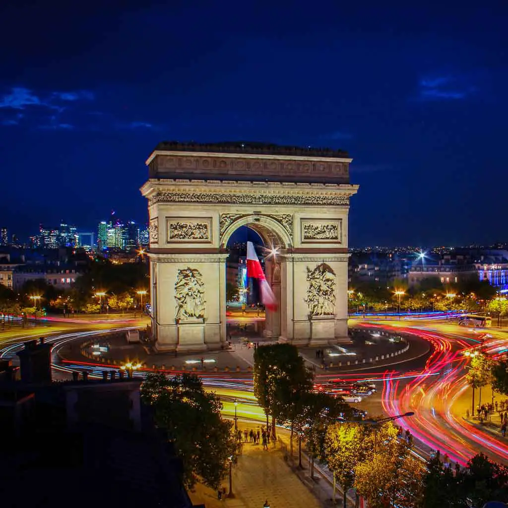 Aerial view of the Arc de Triomphe at night with the flag of France hanging in the middle.