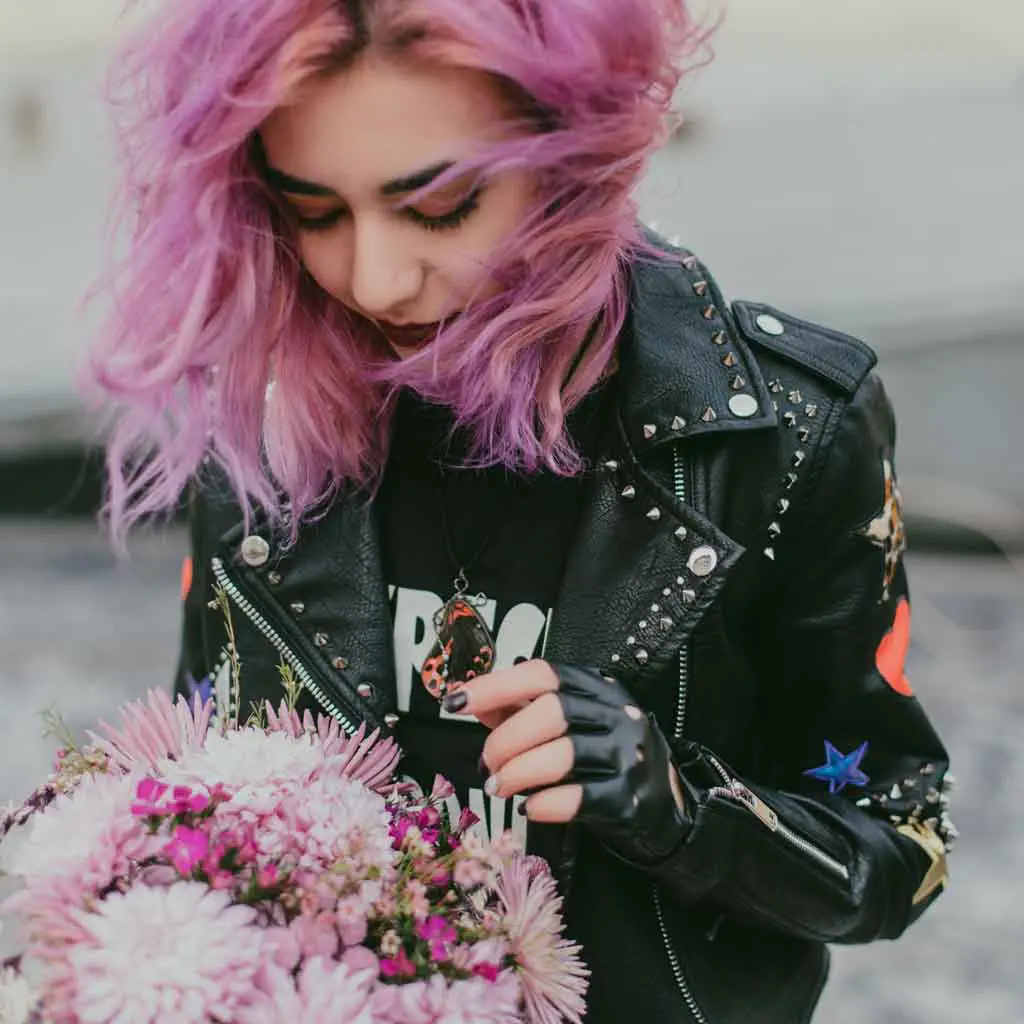 Closeup of a woman with lavender hair wearing a leather jacket and carrying a floral bouquet.