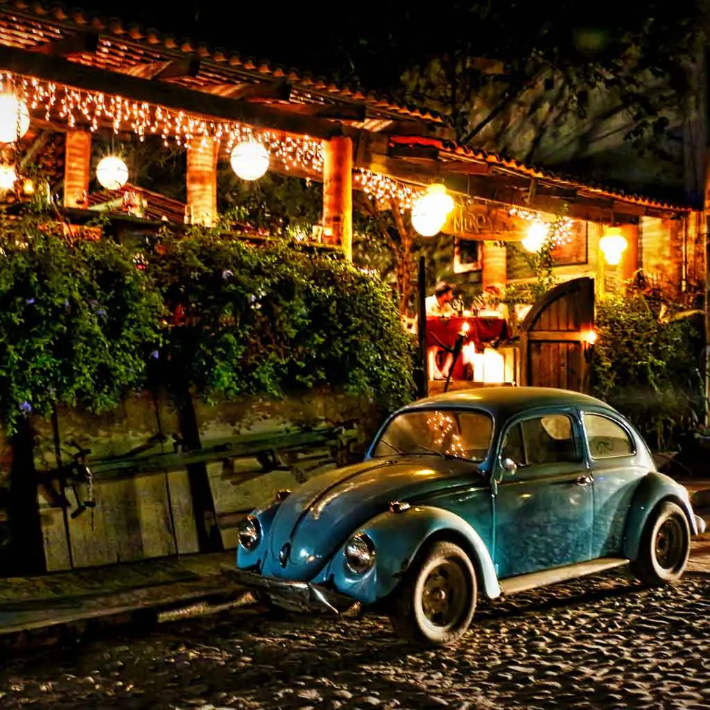 Landscape view of a cobblestone street with a vintage blue Volkswagon beetle parked outside a festive building at night.