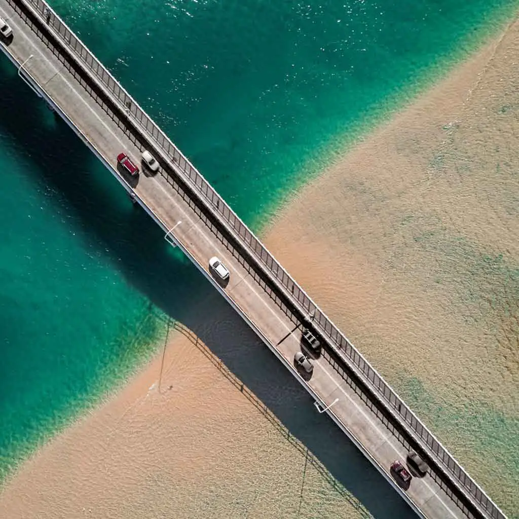 Aerial view of cars driving over a highway over turquoise water and sand.