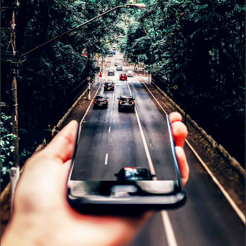 Photo collage of a person holding a phone perpendicular to a road so that it looks like cars are driving out of it onto the road.