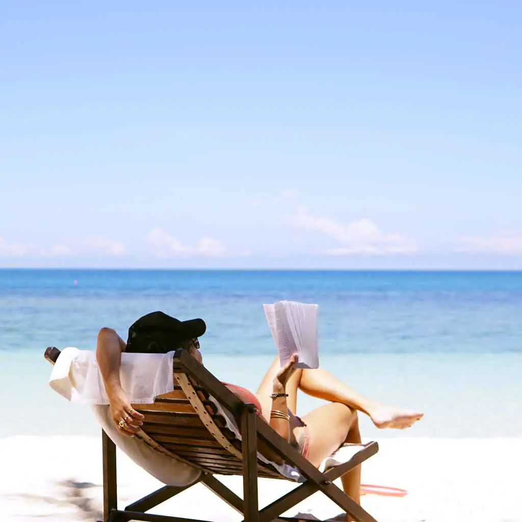 View of a woman from behind in a lounge chair reading a book while sitting on the beach.