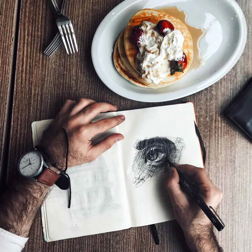 Photo of a man drawing in a sketchbook with a plate of pancakes nearby.