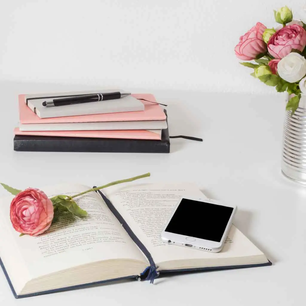 Photo of an open book with an iphone resting on it. Nearby is a stack of journals and a vase of flowers.