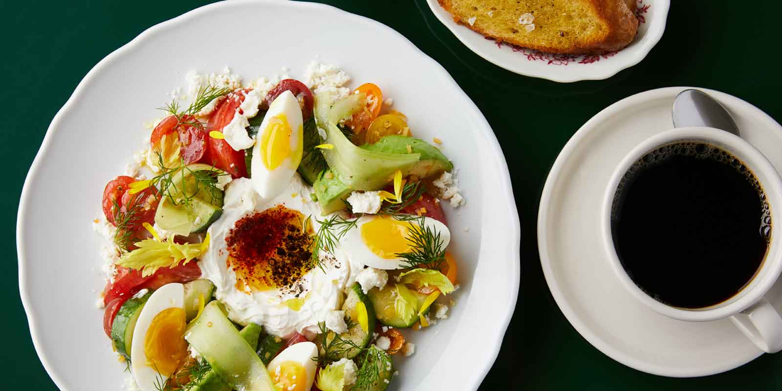 Photo of a plate of veggies, feta cheese, ricotta cheese, and hard boiled eggs next to a cup of black coffee and toast.