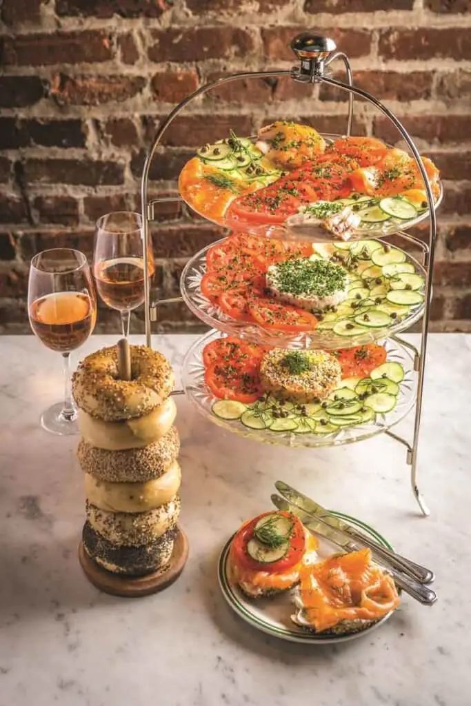 Photo of a tower of bagels next to a tower of vegetables and schmear, alongside 2 glasses of rosé wine.