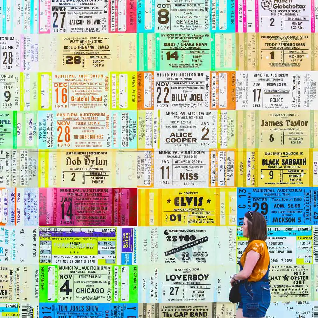 Photo of a woman walking by a wall plastered in giant replicas of concert ticket stubs.