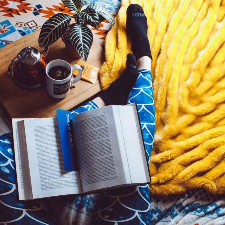 Flatlay photo of a woman's legs with a book on her lap, a large fluffy blanket, and a wooden tray with a cup of tea nearby.