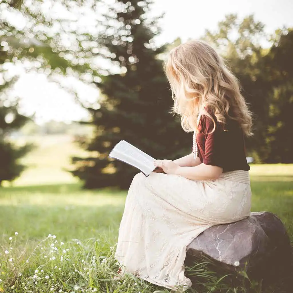 Photo of a woman sitting sideways on a rock while reading a book.