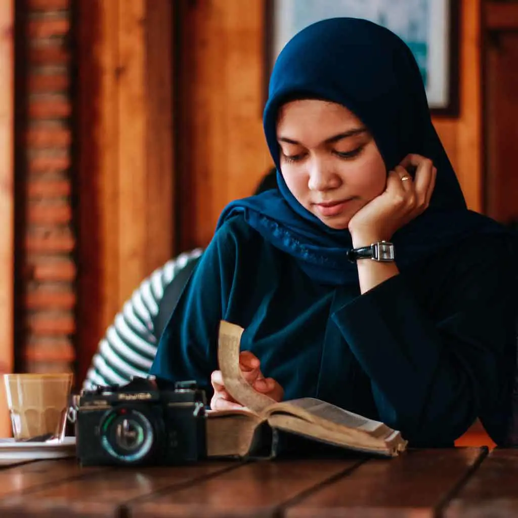 Photo of a woman in a hijab reading a book at a cafe.