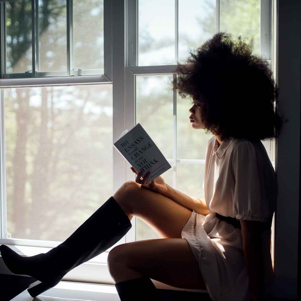 Photo of a woman sitting in a window seat while reading a book.