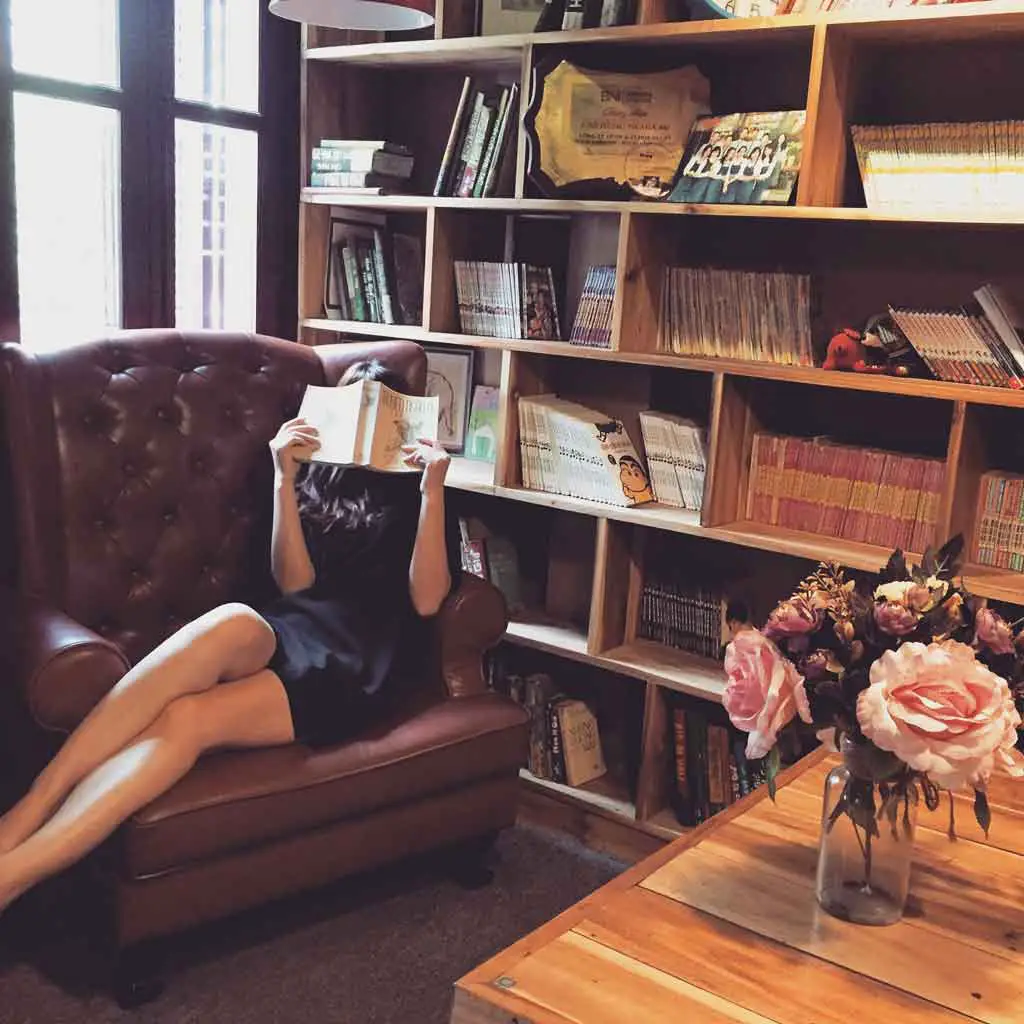 Photo of a woman sitting on a leather chair while reading a book in a library.