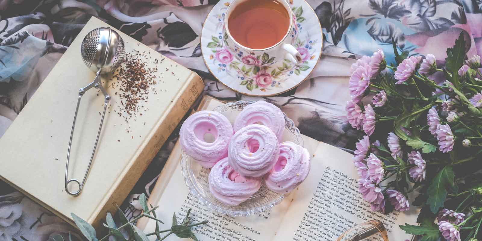 Flatlay photo of old books with a bunch of lavender flowers, pink meringue cookies, and a cup of tea.