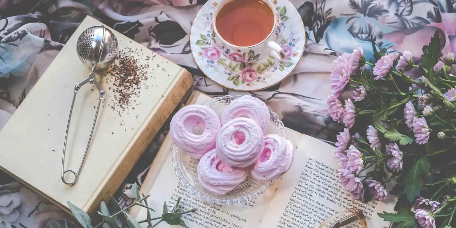 Flatlay photo of old books with a bunch of lavender flowers, pink meringue cookies, and a cup of tea.