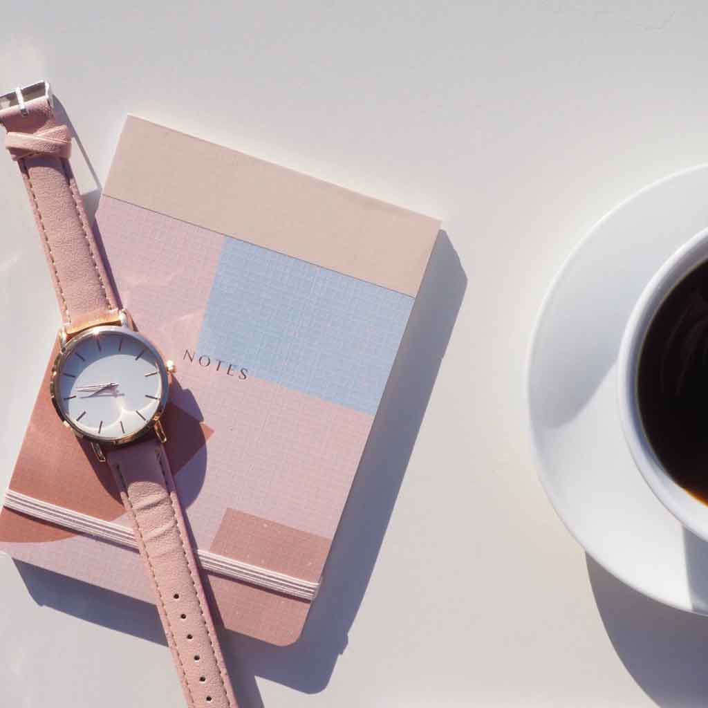 Flat lay photo of a pink and gray notebook on a table with a watch strewn on top and a cup of coffee nearby.