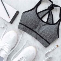 Flat lay photo of a grey and black sports bra, white sneakers, ear buds, measuring tape, and a notepad and pencil.