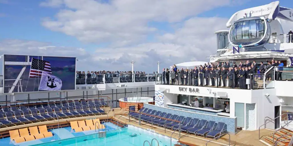 Closeup photo of the dedication ceremony for Royal Caribbean International's Ovation of the Seas cruise ship. People in suits and business attire are standing on the top deck of the ship watching a flag raising ceremony on a large flat screen above the pool area below them.
