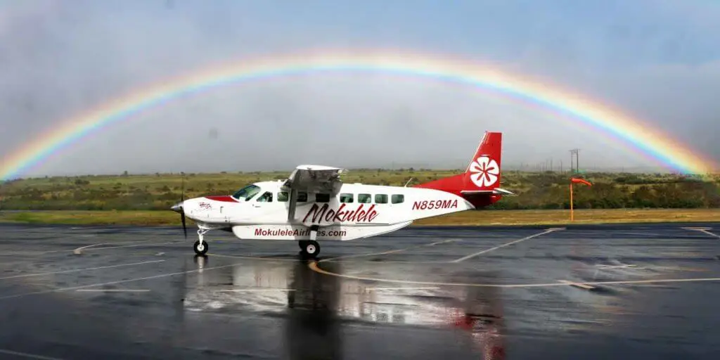 Photo of a Cessna passenger plane at a small airfield with a rainbow overhead. The Plane says Mokulele and MokuleleAirlines.com on the side in the color red.