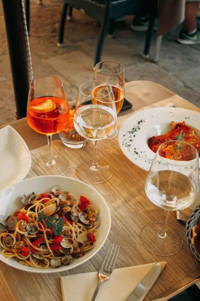 Closeup of a small table with 2 Italian pasta dishes and several wine glasses filled with a drink.