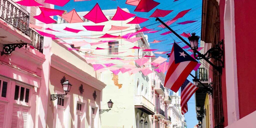 Closeup of pink kites strung up between 2 buildings, with the US and Puerto Rico flags in the distance.