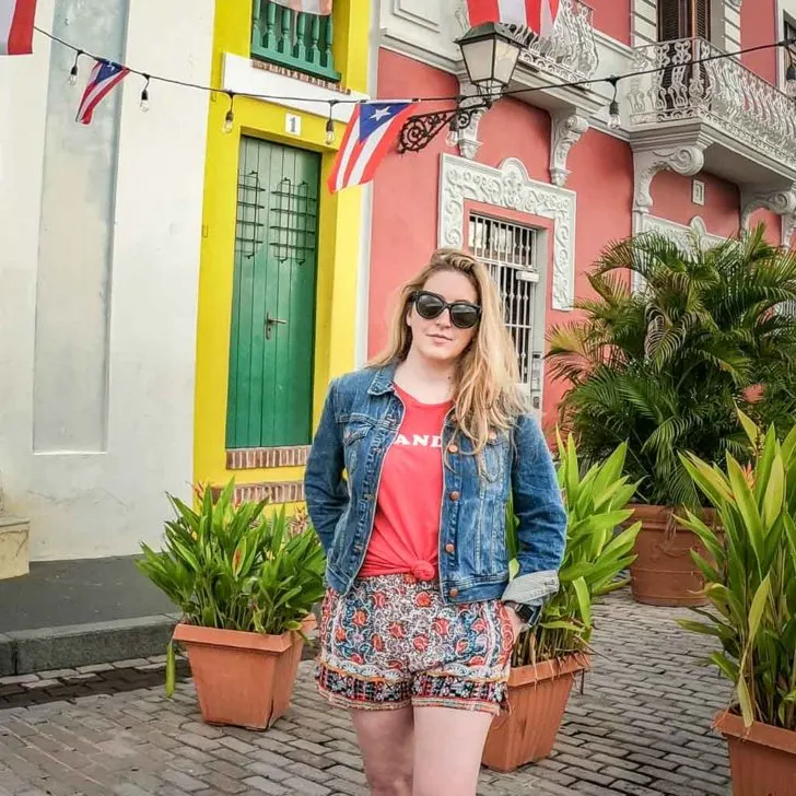 Photo of travel blogger Meg Frost standing in front of colorful buildings with small Puerto Rico flags strung up along a string of lights.