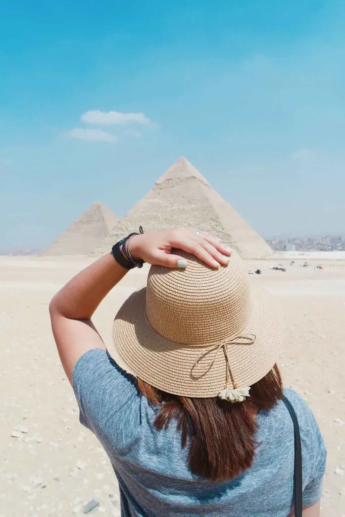 A woman standing with her back to the camera with her hand on her straw hat. In the background are the Great Pyramids of Egypt.