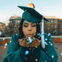 Closeup of a young black woman in dark green graduation cap and gown. She is blowing confetti from her hands toward the camera.