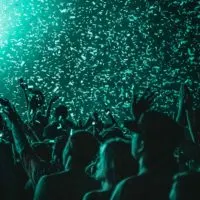 Closeup of a crowd of people in a dark venue with teal lights and confetti falling from above.