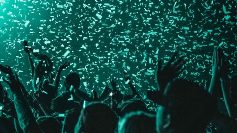 Closeup of a crowd of people in a dark venue with teal lights and confetti falling from above.