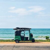 Photo fo a green tuk tuk parked on the side of the road with the ocean in the background.