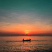 Photo of a gorgeous orange and teal sunset with a silhouette of a small boat with 2 people in it.