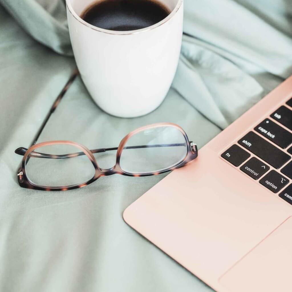 Closeup of a rose gold laptop on top of mint colored sheets with a pair of tortoiseshell glasses and a cup of black coffee next to it.