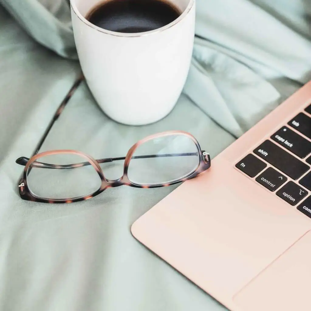 Closeup of a rose gold laptop on top of mint colored sheets with a pair of tortoiseshell glasses and a cup of black coffee next to it.